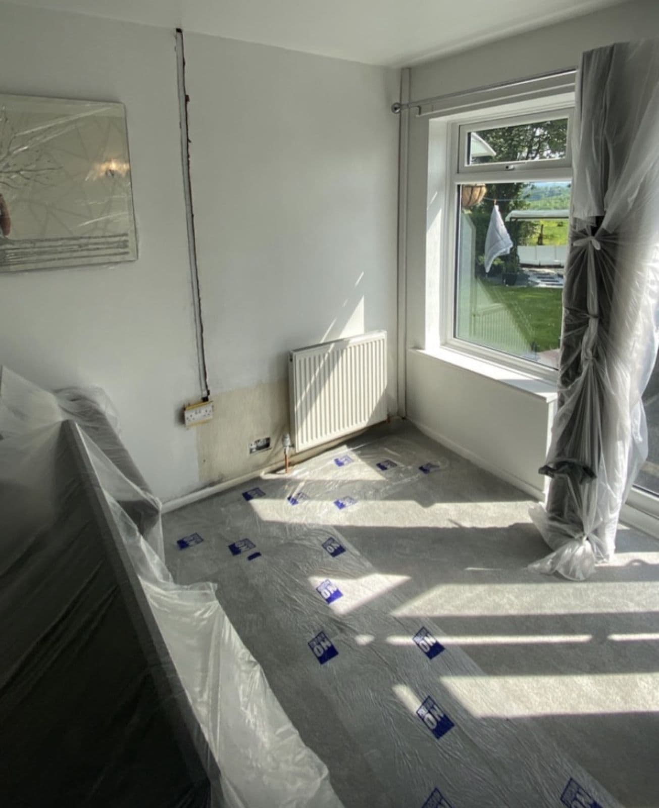 Bed pulled into the middle and sheeted, floor protection down, curtains wrapped so the walls and sockets are free - preparing a home for a house rewire